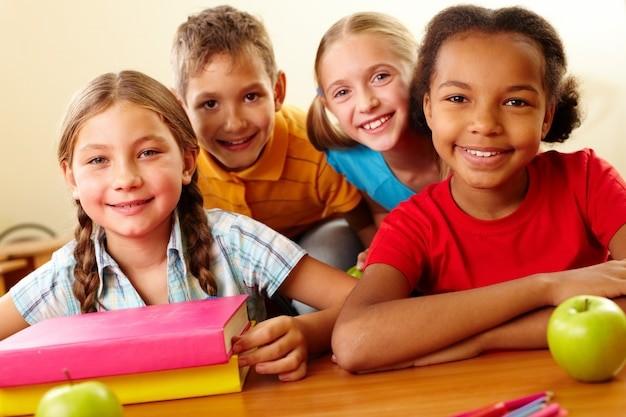 Four children smiling for the camera sitting at a table with books and apples on it 