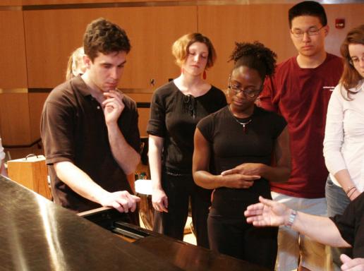 Students stand around a piano a professor is seated at.