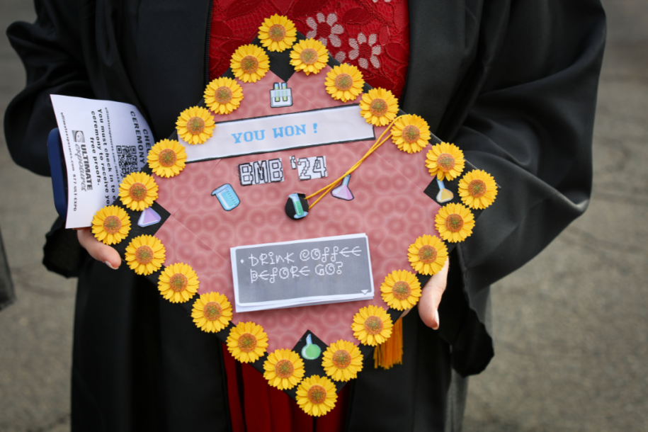 A graduate holds a cap with flower stickers on it ad graphics that say 'you won!' and 'drink coffee before you go?'