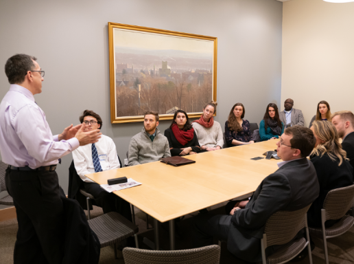 A group of students sit at a conference room table while a lecturer speaks.